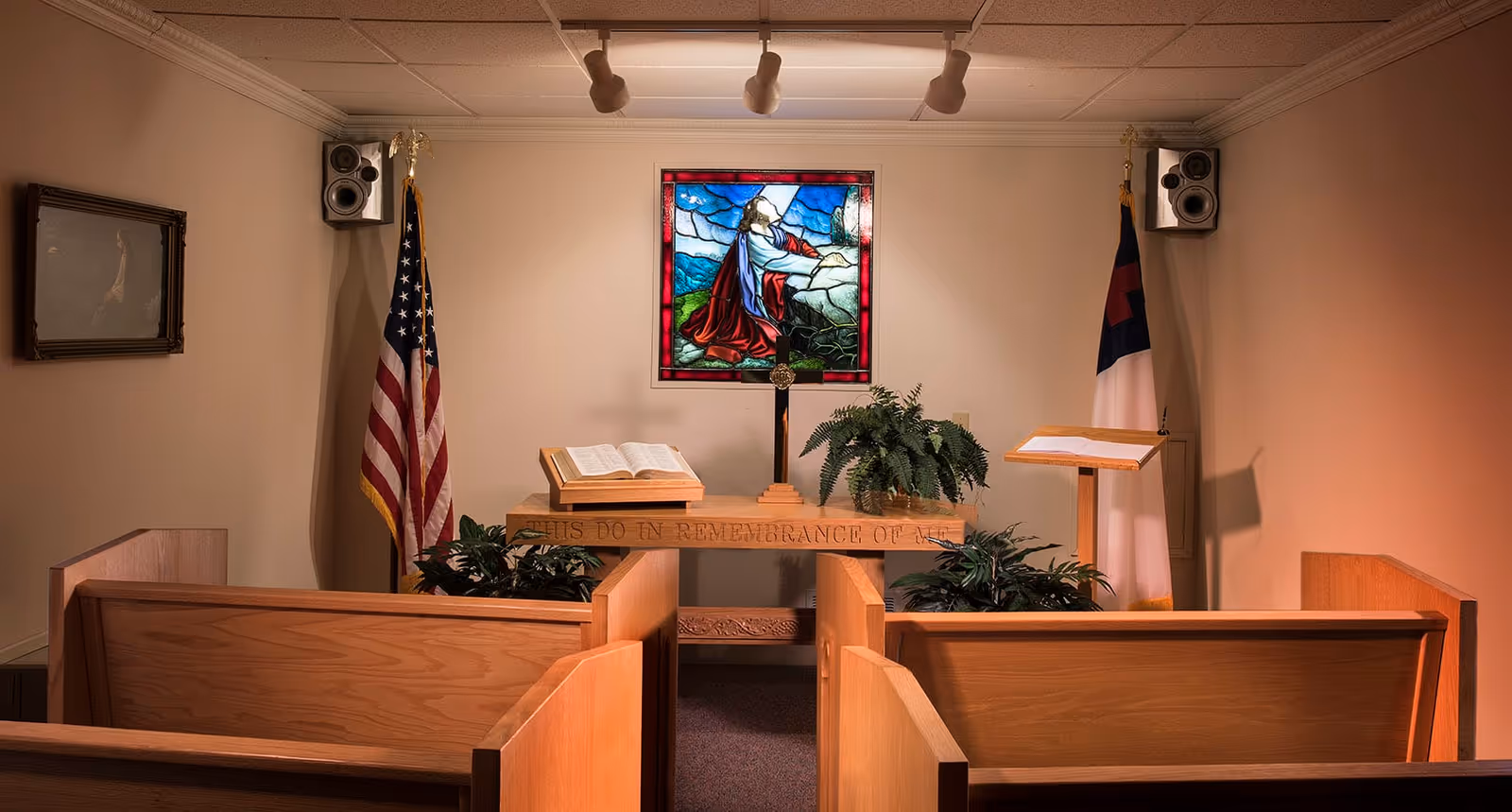 Small chapel room with wooden pews facing a wooden altar. On the altar is an open Bible, a cross, and a plant. Behind the altar is a stained glass window depicting a religious figure kneeling in prayer. Two flags, an American flag and a Christian flag, stand on either side of the altar. Speakers are mounted on the upper corners of the walls.