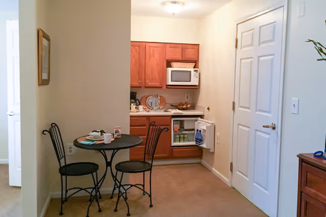 Small studio kitchenette with a round bistro table and two chairs, wood cabinets, microwave, and an open mini-fridge.