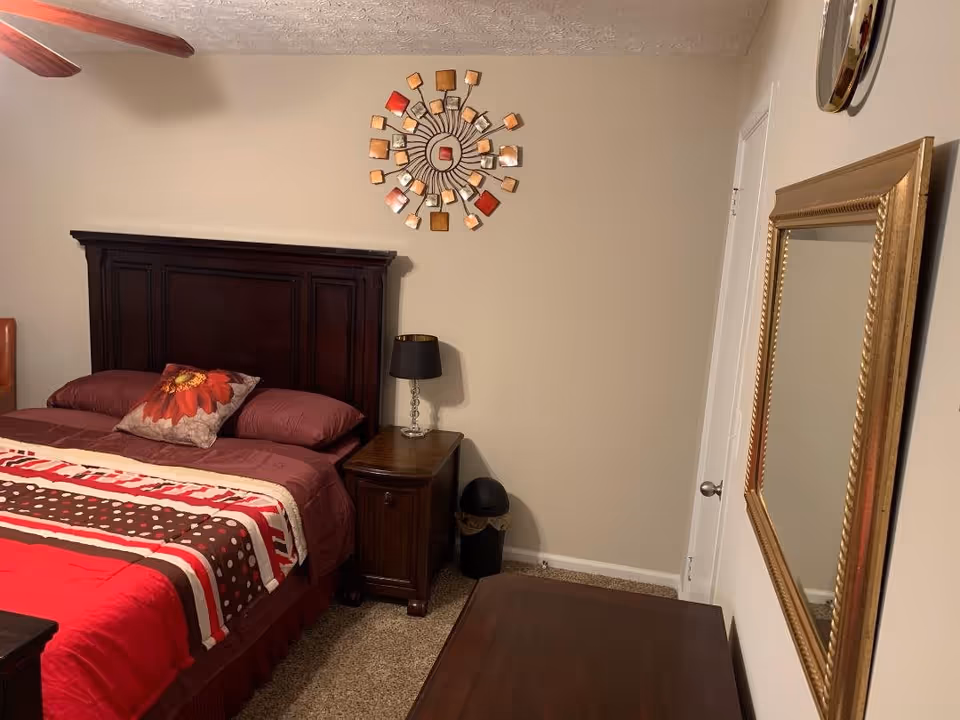 A bedroom with a dark wooden bed frame and matching nightstand. The bed is made with red, brown, and white bedding and a decorative pillow with a flower design. A black lamp with a clear base sits on the nightstand. Above the bed on the beige wall is a decorative metal wall art piece with square and rectangular shapes in shades of orange, red, and brown. There is a large gold-framed mirror on the adjacent wall, and a ceiling fan with wooden blades is partially visible.