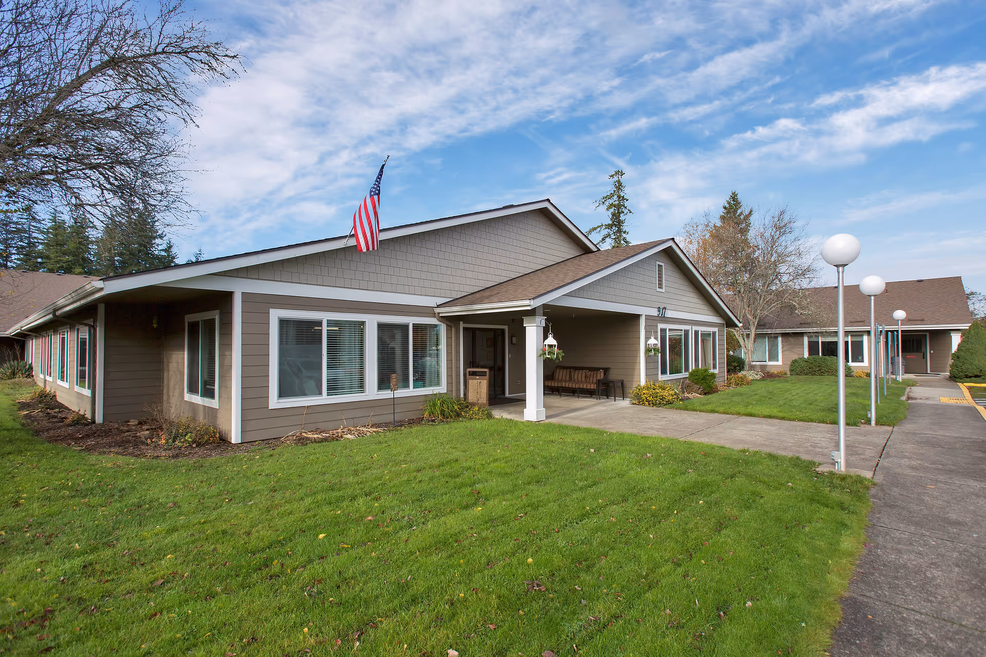 Single-story senior living facility building with an American flag, covered entrance, and a grassy lawn under a partly cloudy sky.