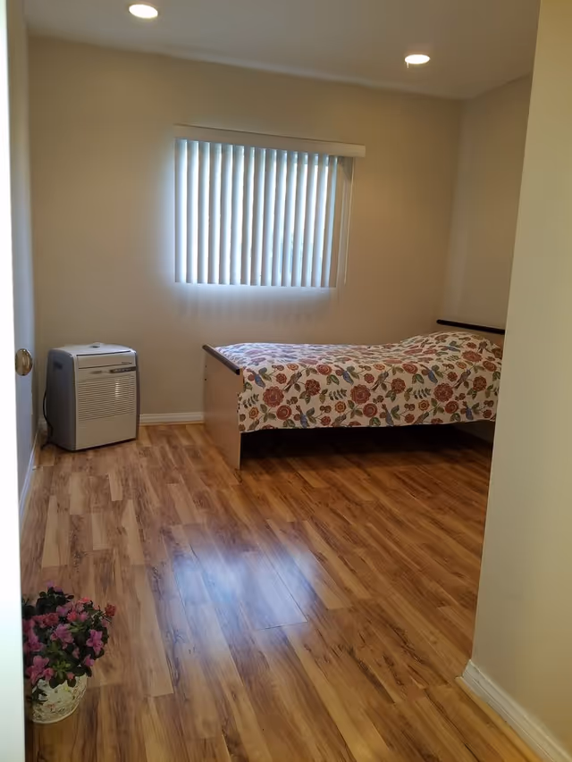 A simple bedroom with wooden flooring, a single bed covered with a floral patterned bedspread, a window with vertical blinds, a small air purifier or heater in the corner, and a potted plant with pink flowers on the floor near the entrance.