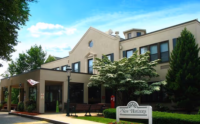 Front exterior of the New Horizons senior living building showing the entrance canopy, benches, landscaping, and a sign that reads "New Horizons".