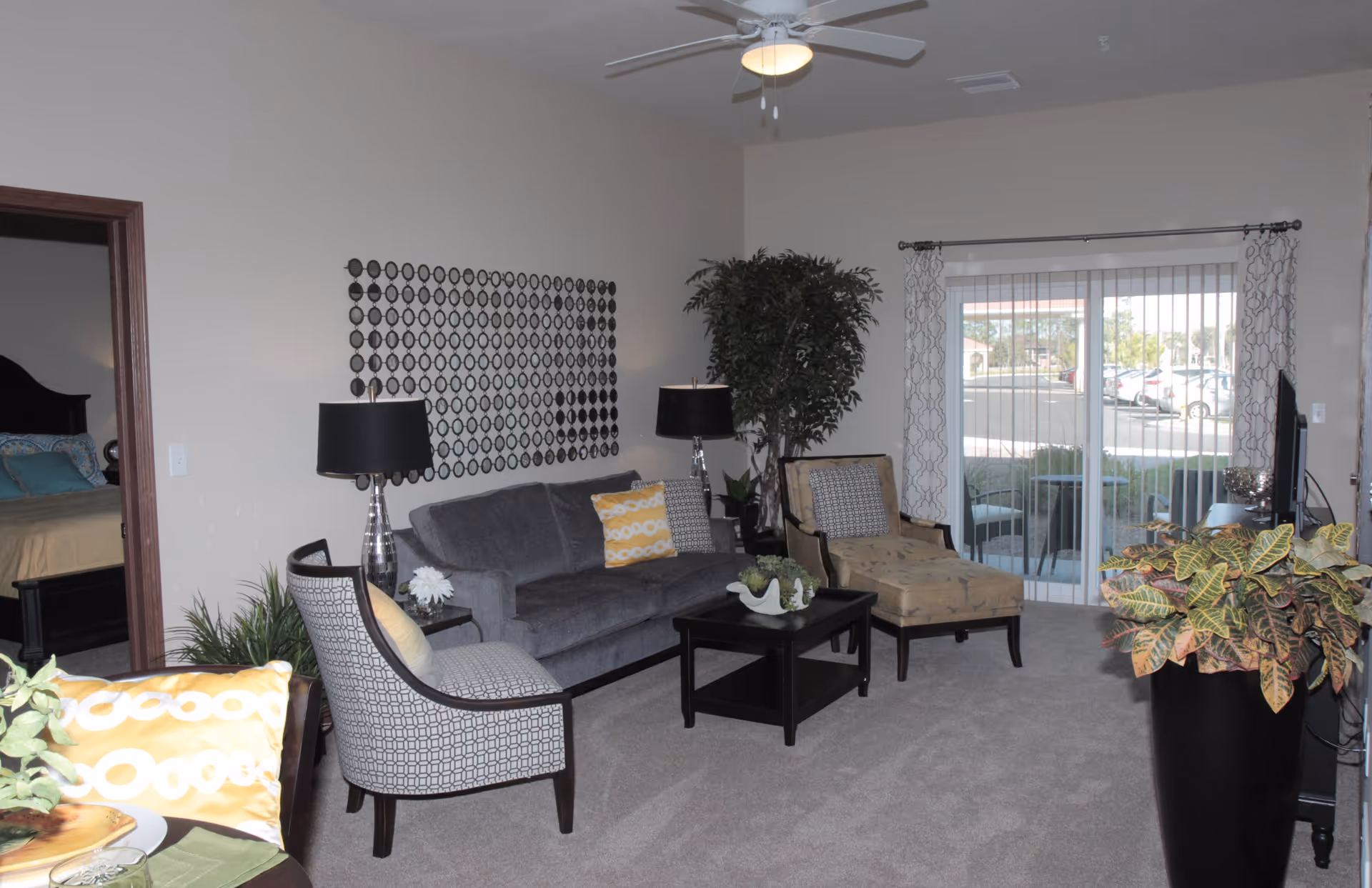 A cozy living room in a retirement community featuring a gray sofa with yellow and patterned pillows, two armchairs, a black coffee table with decorative items, a floor lamp, a large potted plant, and a sliding glass door with vertical blinds leading to an outdoor area. A bedroom is partially visible through an open doorway on the left.