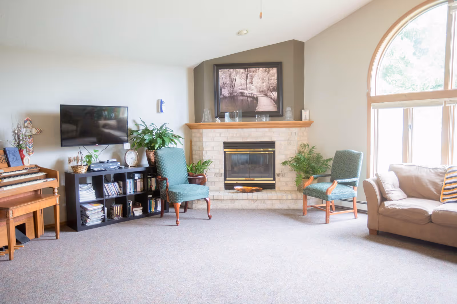 Bright communal living room with a fireplace, TV and bookshelf, two upholstered chairs, a sofa, and a large arched window.