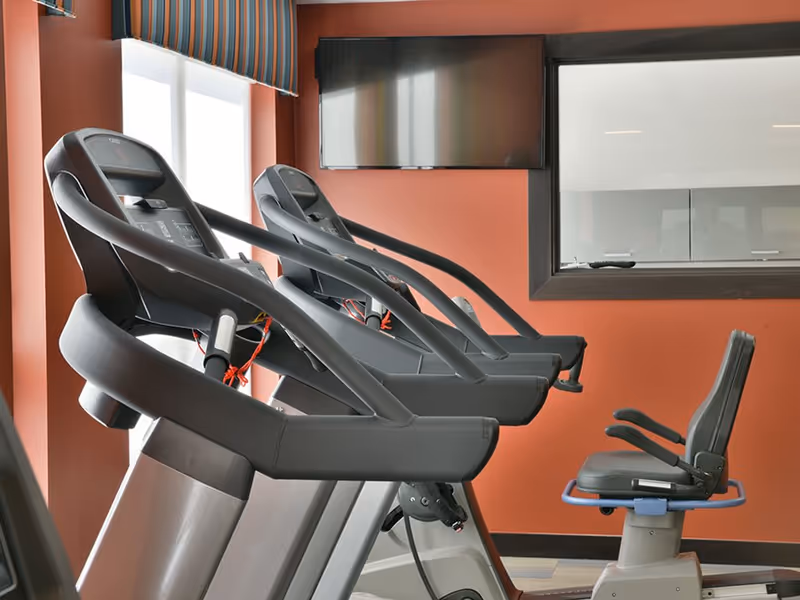 A fitness room with several treadmills and a recumbent exercise bike positioned in front of a wall-mounted flat screen TV. The walls are painted a warm orange color and there is a window with a striped valance.