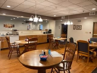 A spacious dining area in an assisted living facility featuring several wooden tables and chairs arranged neatly. The room has warm lighting with hanging light fixtures, a wooden floor, and a counter area in the background with kitchen appliances and cabinets. The walls are decorated with framed pictures.