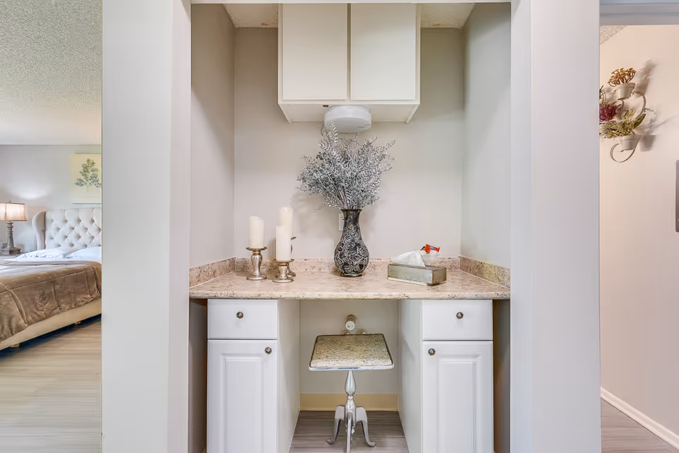A small vanity area with a marble countertop, white cabinets, and a decorative vase with silver foliage. There are three candles on silver holders and a tissue box on the countertop. A small stool is tucked under the counter. To the left, a bedroom with a tufted headboard and a bedside lamp is partially visible.