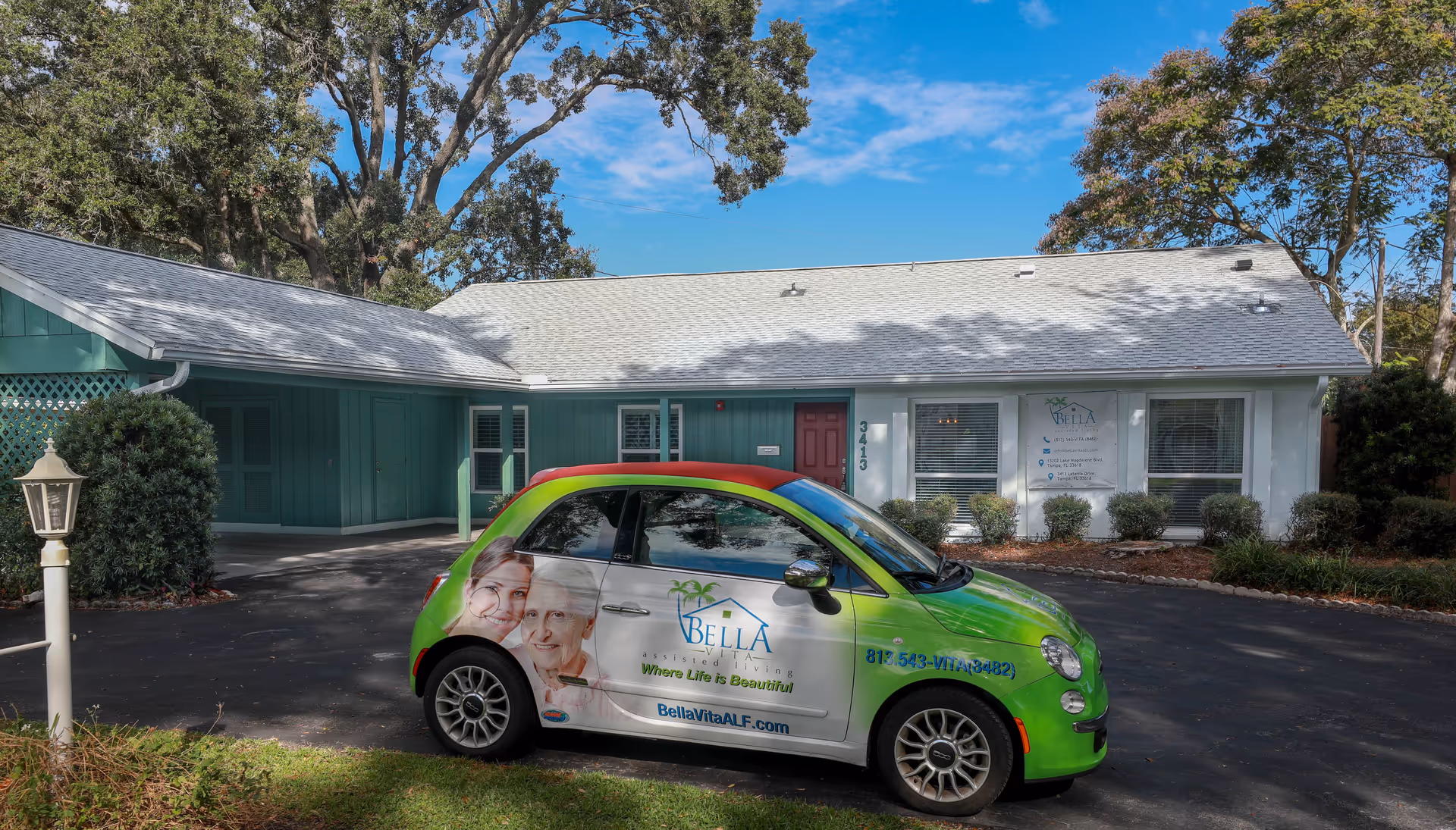 A small green and white car with Bella Vita ALF branding parked in front of a single-story building with teal and white exterior walls, a red door, and several windows. The building is surrounded by trees and bushes under a blue sky with some clouds.