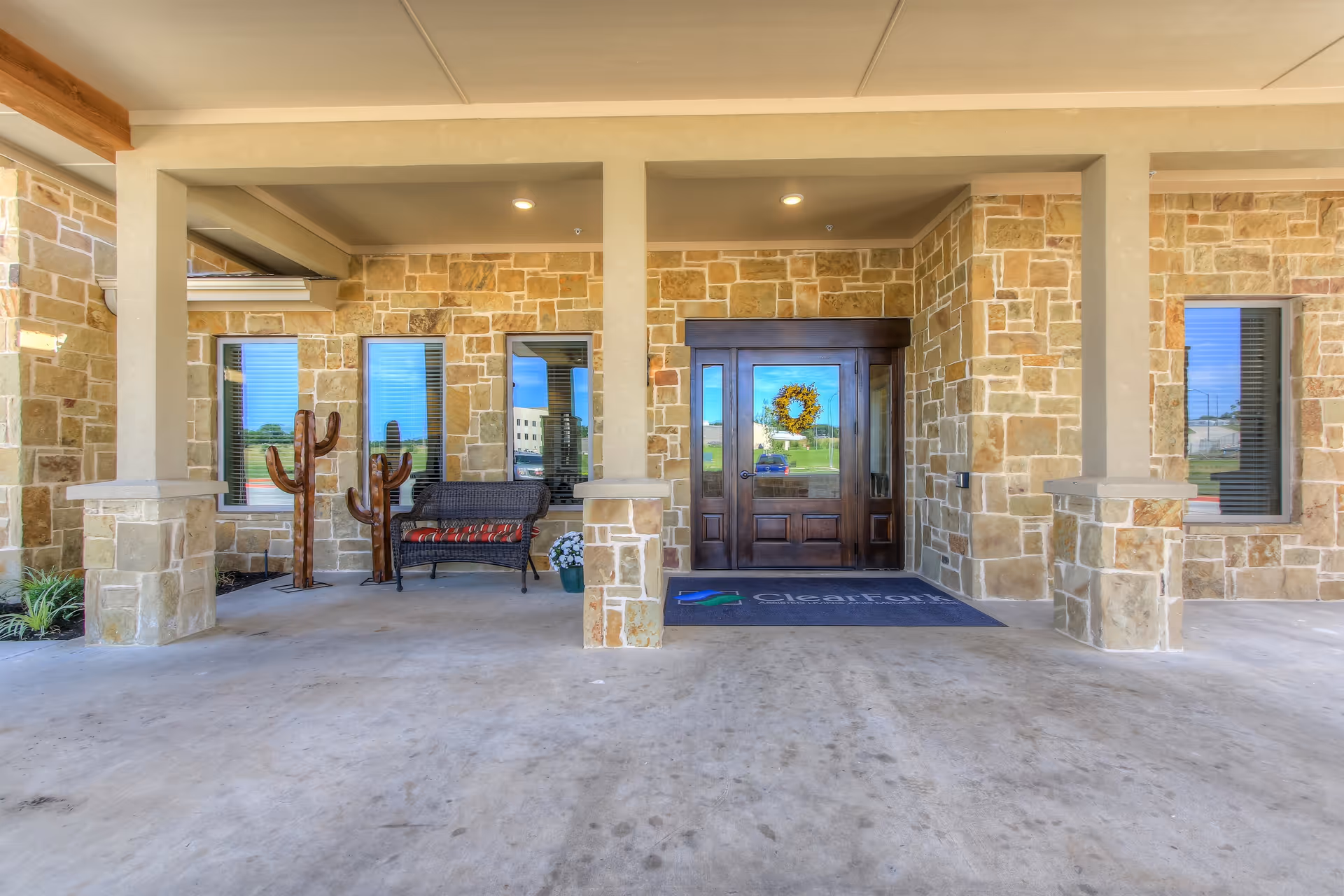Entrance area of Clear Fork Assisted Living and Memory Care facility with stone walls, a wooden door decorated with a wreath, two windows, a bench with a cushion, and two decorative wooden cactus sculptures.