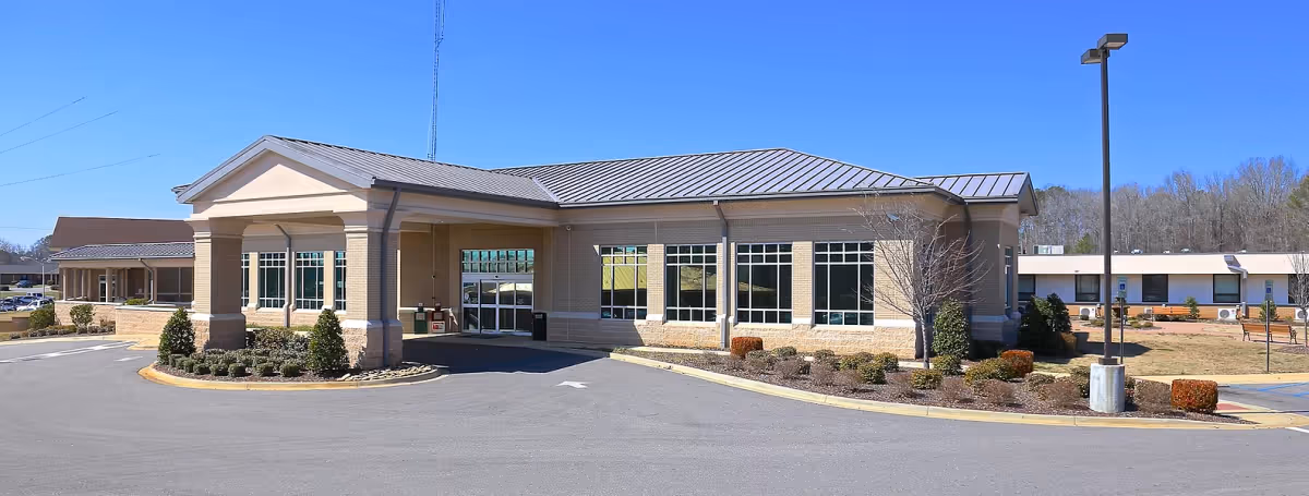 Exterior view of Bibb Med Center Nursing Home building with a covered entrance, large windows, landscaped bushes, and a clear blue sky.