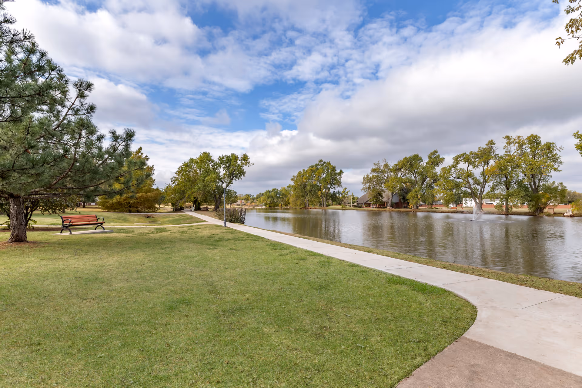 A peaceful outdoor scene featuring a paved walking path alongside a calm pond with a fountain. The area is surrounded by green grass, trees, and a wooden bench under a partly cloudy sky.