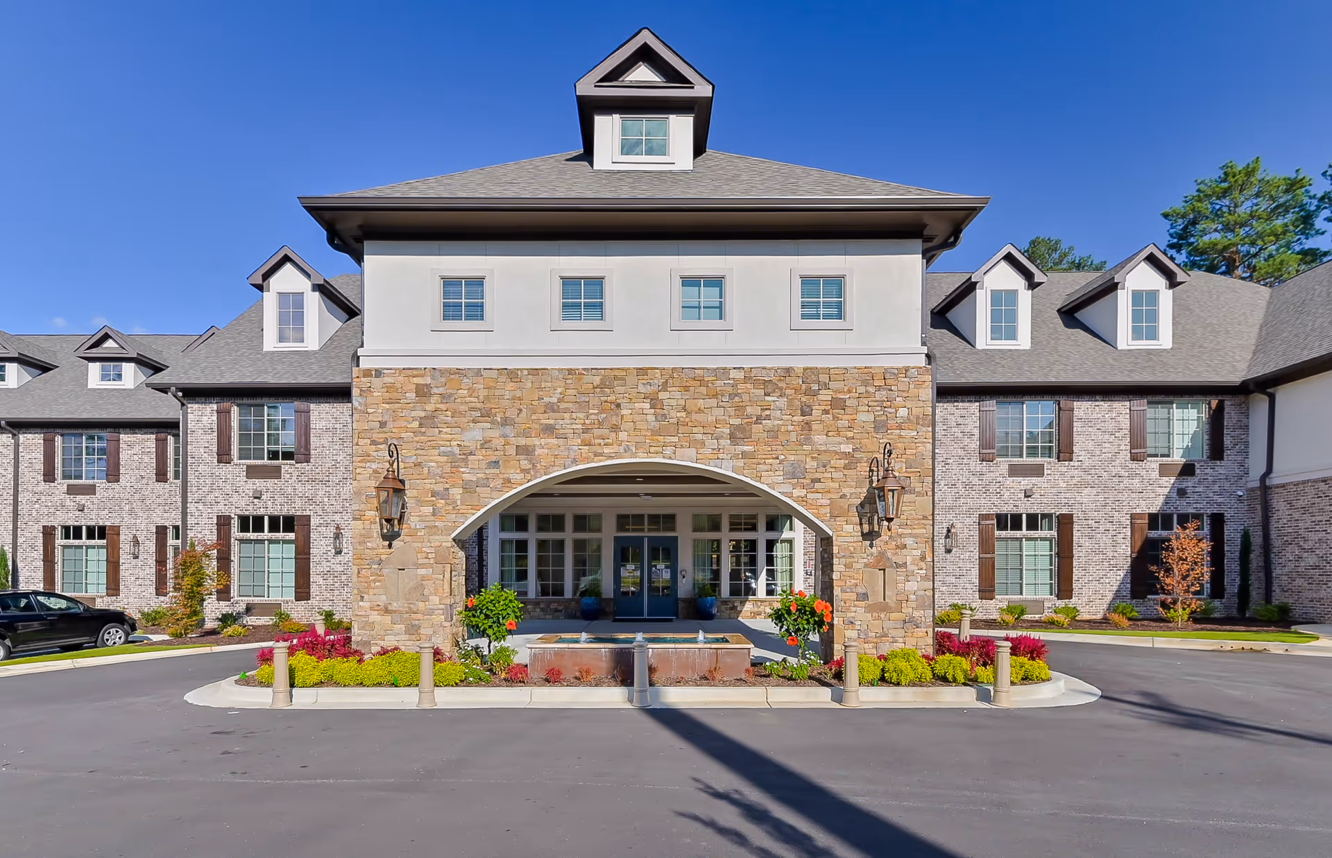 Front exterior view of Orchard at Brookhaven facility featuring a large stone and brick building with multiple windows, a central archway entrance, decorative lanterns, and landscaped flower beds under a clear blue sky.