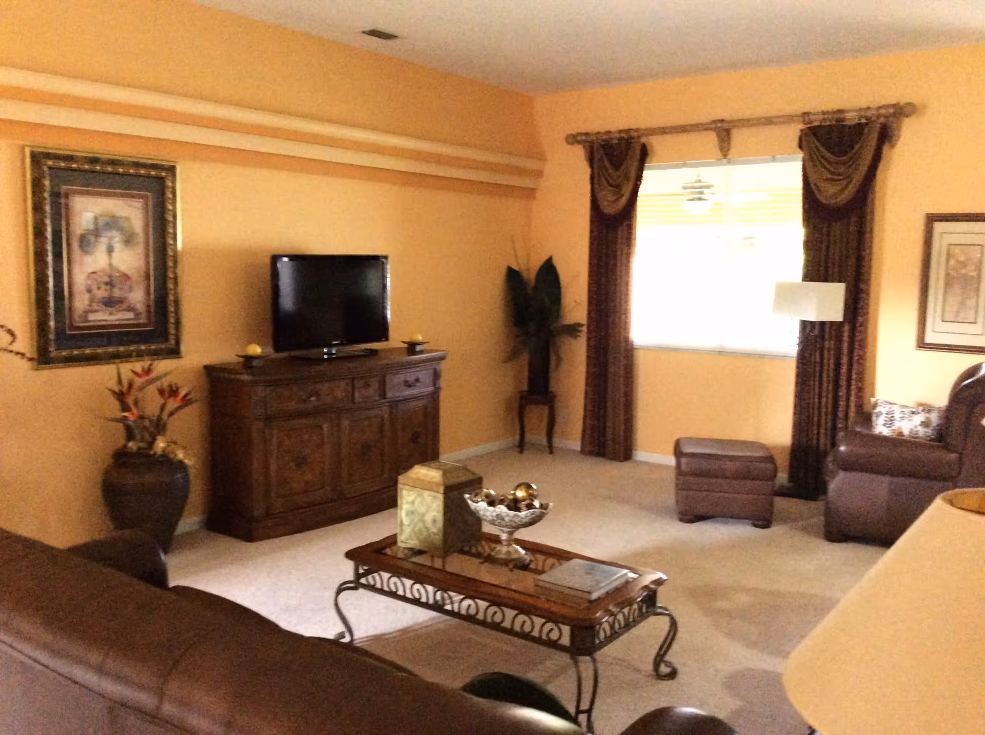 A cozy living room with peach-colored walls, a brown leather sofa, a matching ottoman, a wooden coffee table with decorative items, a wooden cabinet with a flat-screen TV on top, framed artwork on the walls, a window with brown curtains, and a floor lamp.