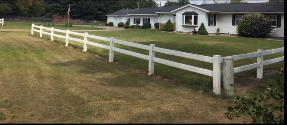 A single-story white building with a dark roof behind a white wooden fence, surrounded by a grassy lawn and some bushes and trees in the background.