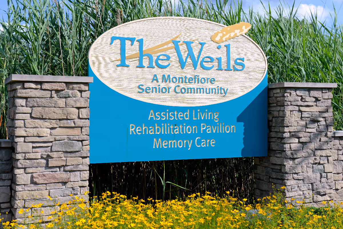 A large outdoor sign for The Weils, a Montefiore Senior Community, indicating services including Assisted Living, Rehabilitation Pavilion, and Memory Care. The sign is mounted between two stone pillars with tall green plants and yellow flowers in the foreground.