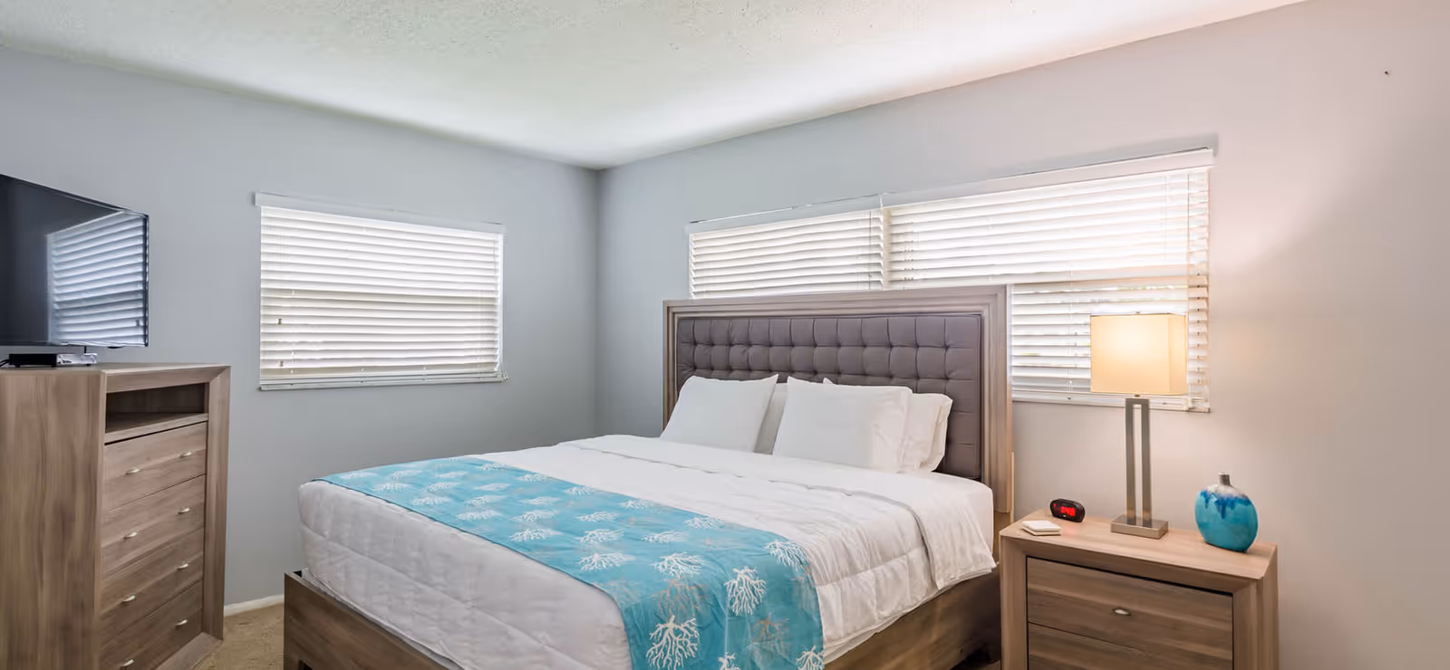 A bright bedroom with a large bed featuring a tufted headboard, white bedding, and a light blue throw blanket with a coral pattern. The room has two windows with white blinds, a wooden dresser with a flat-screen TV on top, and a wooden nightstand with a lamp, a small blue vase, and a digital alarm clock.