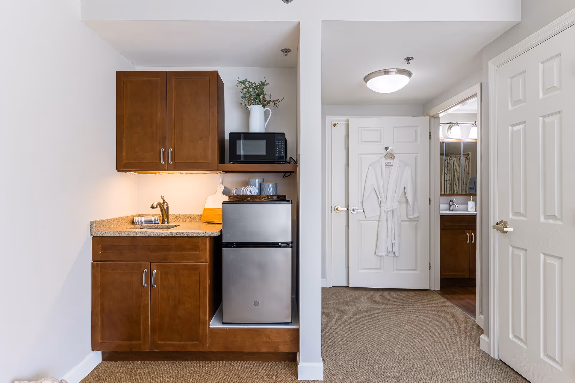 A small kitchenette area with wooden cabinets, a granite countertop with a sink, a mini refrigerator, and a microwave. A white pitcher with greenery is placed on the microwave. To the right, there is a white door with a white robe hanging on it, and an open door leading to a bathroom with a sink and mirror visible.
