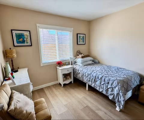 A small, cozy bedroom with a single bed covered in a gray quilted blanket and several pillows. Next to the bed is a white nightstand with a small lamp, a photo frame, and a flower vase. There is a window with white blinds on the wall behind the bed. On the left side of the room, there is a beige armchair with a pillow and a table with a lamp and decorative items. The walls are painted light beige and the floor is wooden.