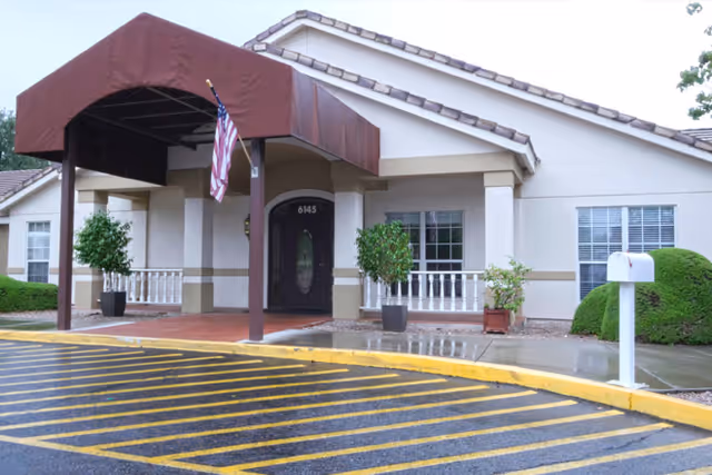 Entrance of a single-story building with a covered porch supported by columns, an American flag hanging from the porch, two potted plants on either side of the door, and a white mailbox on the right. The building has beige walls, a tiled roof, and several windows with white blinds. The ground is wet and there are yellow-striped parking or no-parking zones in front of the entrance.