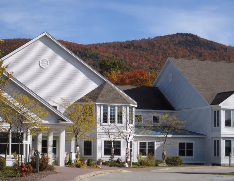 Exterior view of a white senior living facility building with multiple windows and peaked roofs, set against a backdrop of a forested mountain with autumn foliage under a clear blue sky.