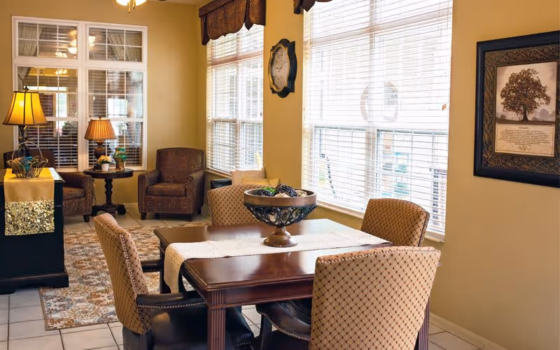 A cozy interior room with a wooden dining table surrounded by four upholstered chairs. The table has a decorative bowl centerpiece and a runner. In the background, there are two armchairs with a small round table between them, holding a lamp and a plant. Large windows with blinds allow natural light to fill the room. The walls are painted a warm beige color and decorated with a framed picture and a clock.