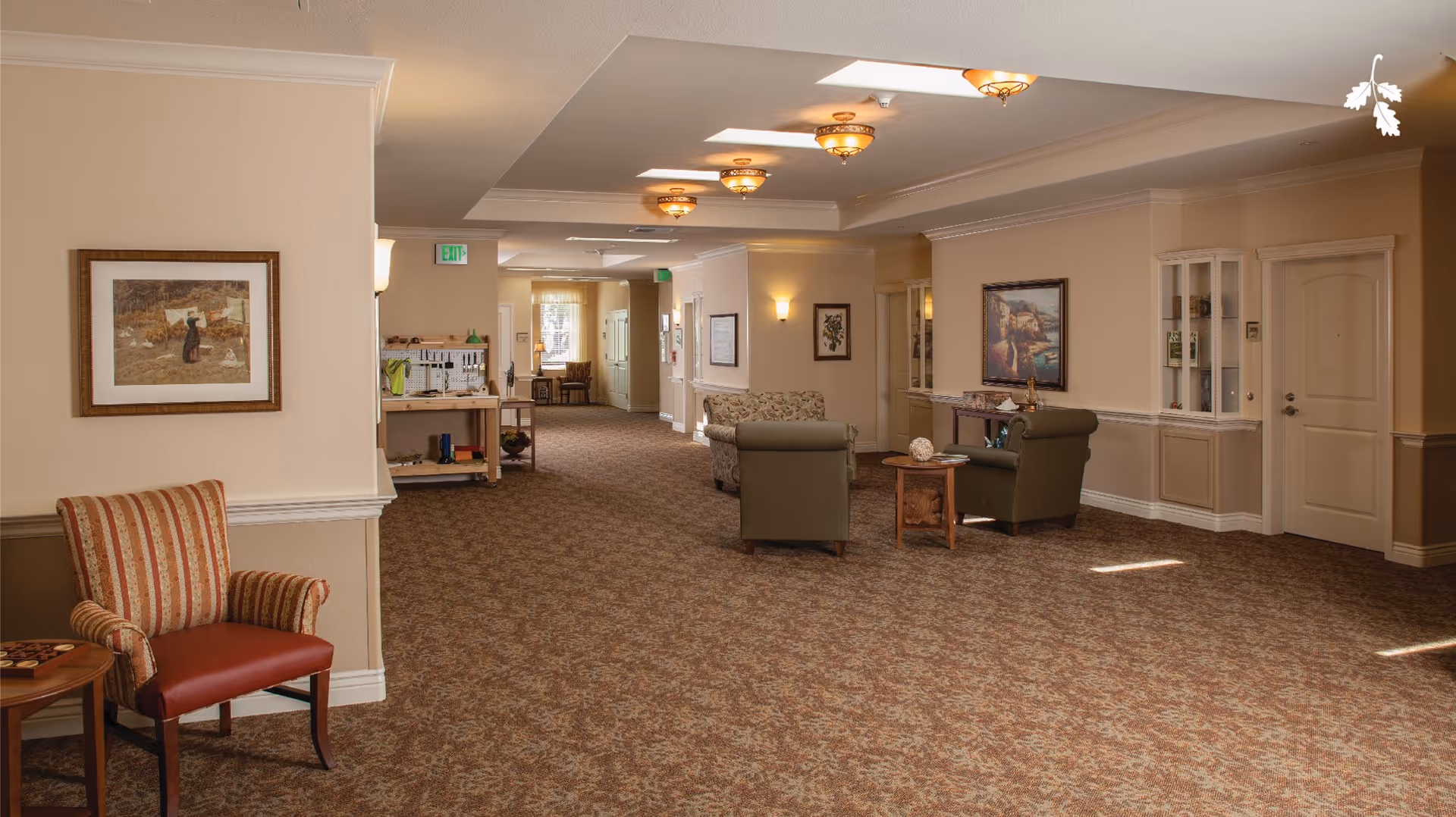 A spacious, carpeted hallway in a memory care community with beige walls and ceiling lights. The hallway features several seating areas with armchairs and small tables, framed artwork on the walls, and a workbench with tools in the background near a window.
