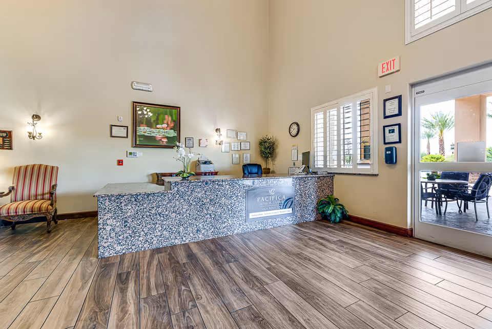 Reception area of San Martin Senior Living with a granite front desk, a striped armchair to the left, framed artwork and certificates on the wall, a clock, and a glass door leading outside to a patio with chairs and palm trees visible.