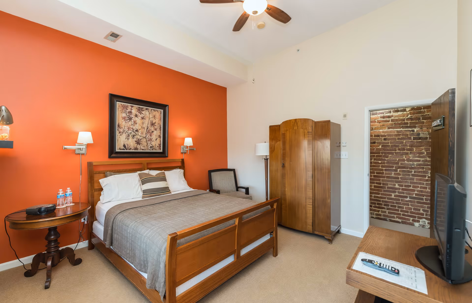 Well-lit bedroom with a wooden bed against an orange accent wall, nightstand, wardrobe, chair, and a TV on a dresser.