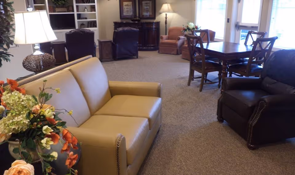 A cozy living room area with a beige leather sofa, a dark brown leather armchair, a wooden dining table with four chairs, and additional seating including two brown chairs and a peach armchair. The room is carpeted and decorated with a table lamp, flowers, and framed artwork on the walls. Large windows allow natural light to fill the space.