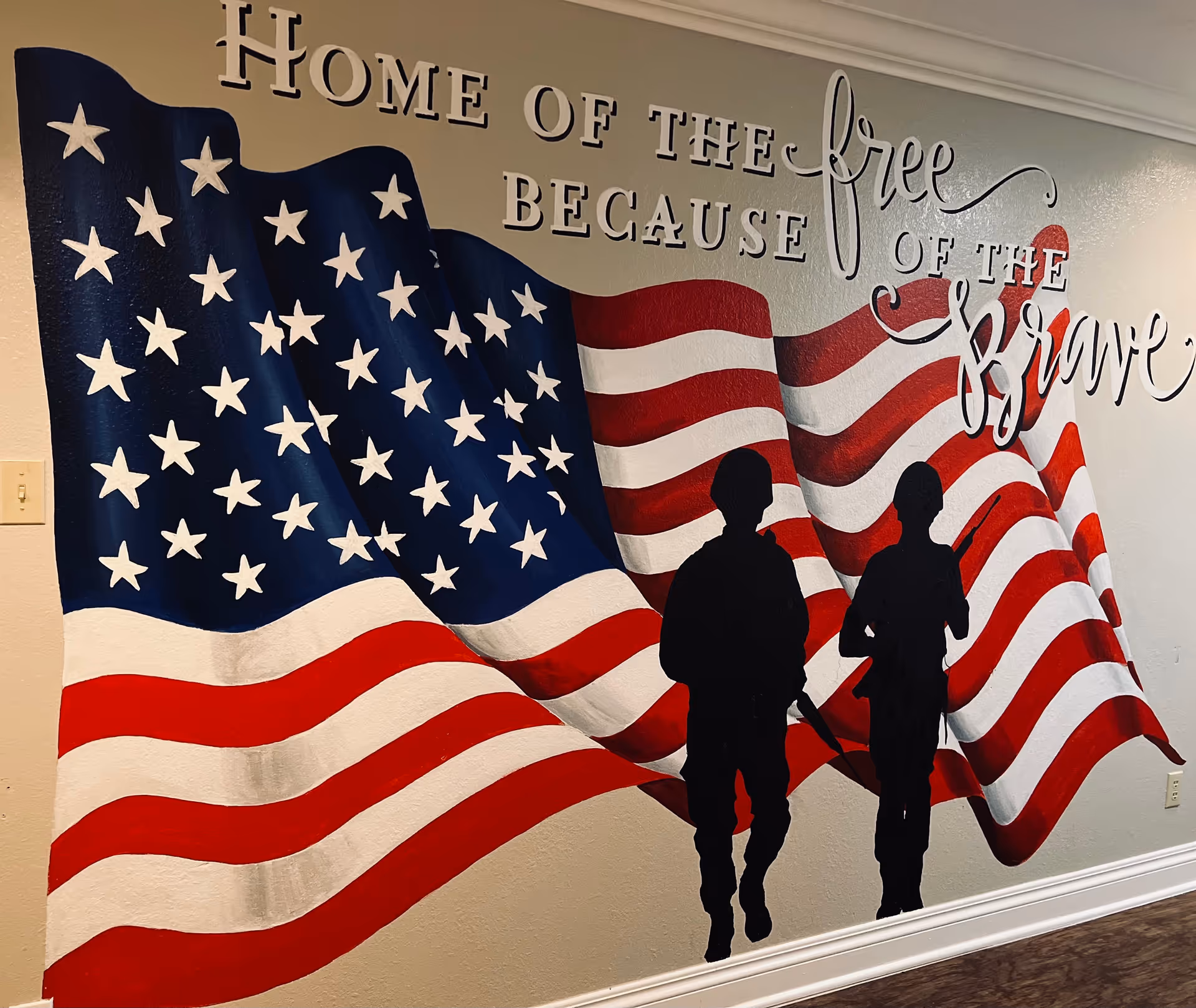A mural painted on a wall featuring a large American flag with two black silhouettes of soldiers in front. Above the flag, the text reads 'Home of the free because of the brave' in a combination of block and cursive lettering.