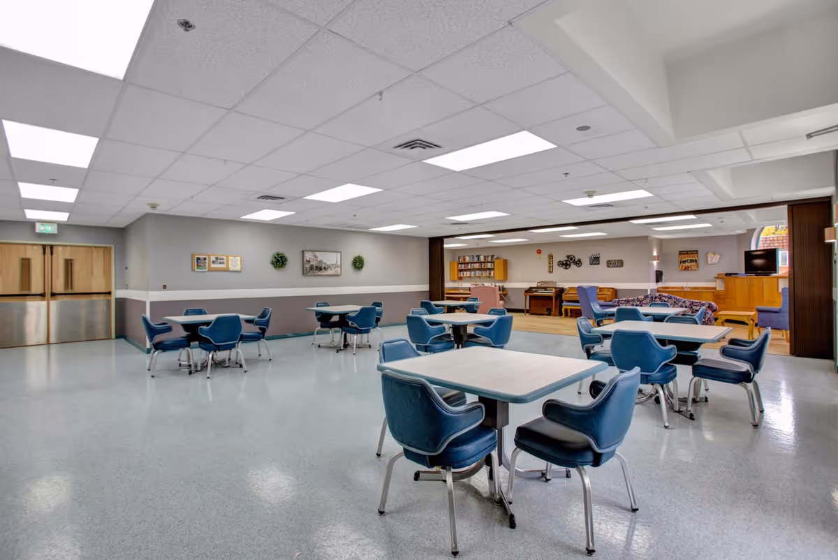A spacious common area in a senior living facility with multiple square tables surrounded by blue cushioned chairs. The room has a light gray floor, white ceiling with fluorescent lights, and light gray walls with a white trim. In the background, there is a cozy lounge area with bookshelves, a piano, and additional seating including a floral couch and purple armchairs. The room appears clean and well-lit.