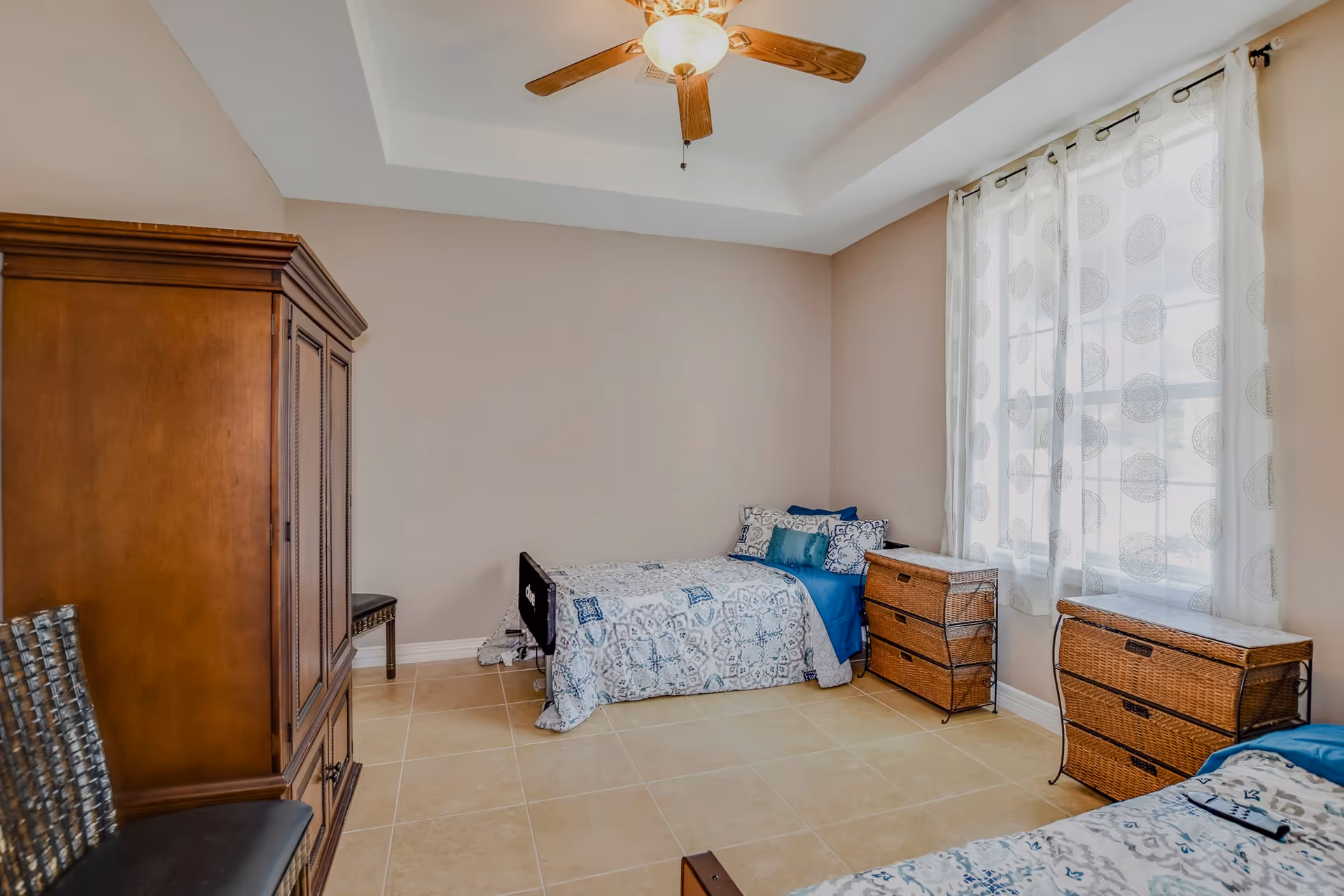 Bright bedroom with two twin beds, a wooden armoire, wicker dressers, a ceiling fan, and a large curtained window.
