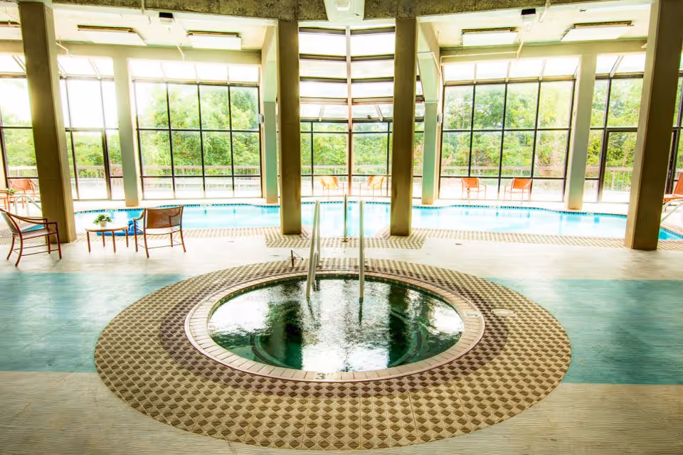 Indoor pool area with a circular hot tub in the foreground surrounded by patterned tiles. Large floor-to-ceiling windows in the background let in natural light and show greenery outside. Several chairs and small tables are placed around the pool area.