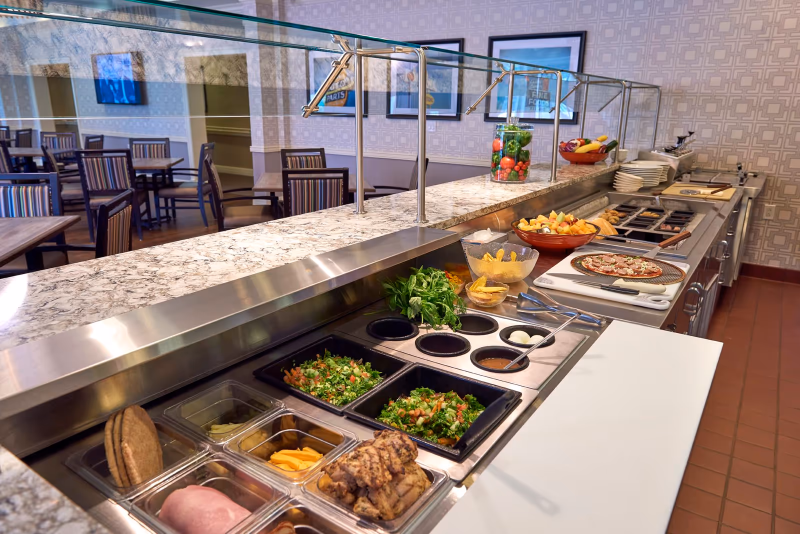 A buffet-style food serving area with a variety of fresh ingredients including salad, sliced meats, cheese, boiled eggs, fruit, and a prepared pizza. Behind the counter, there are tables and chairs in a dining area with framed pictures on the wall.