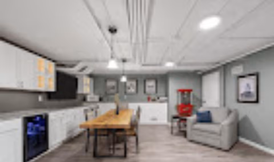 A modern communal kitchen and dining area with white cabinets, a wooden dining table with chairs, a gray armchair with a blue pillow, and a red popcorn machine. The room has light gray walls, framed pictures, and ceiling lights.