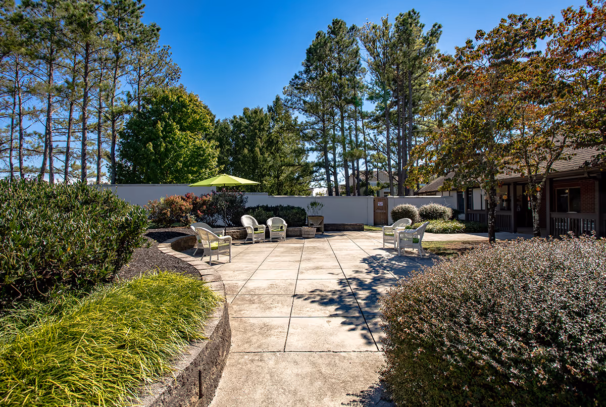 Sunlit outdoor courtyard at a senior living facility with a paved patio, chairs, landscaping, and tall trees.