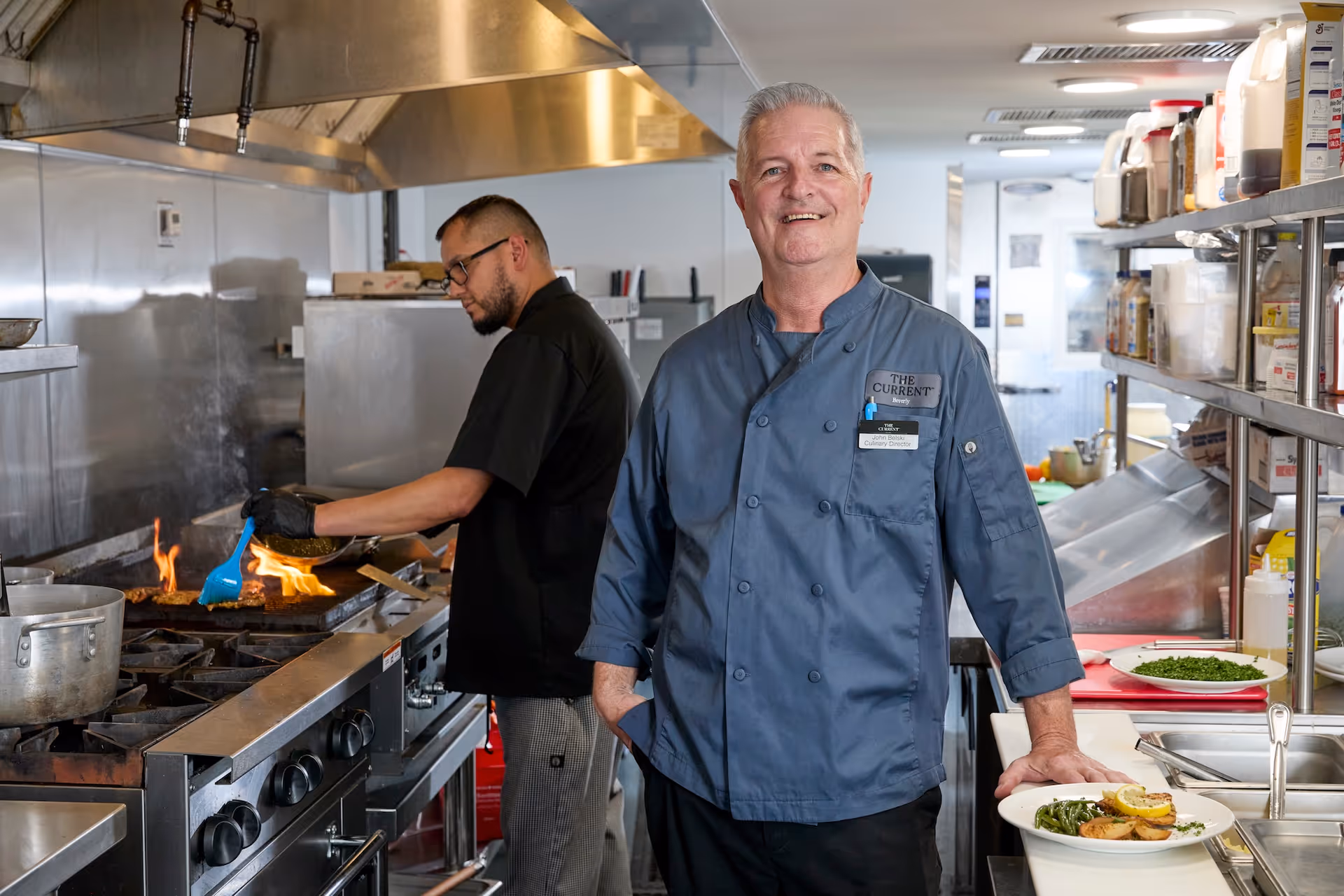 Two chefs in a commercial kitchen. One chef in a black uniform is cooking on a stove with flames visible, while the other chef in a blue uniform with a name tag that reads 'The Current Beverly, John Belski, Culinary Director' is smiling and standing next to a counter with a plated dish of food.