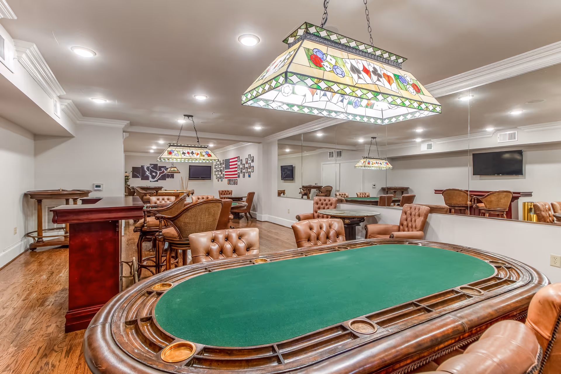 Wood-floored game room with a green-felt poker table, tufted leather chairs, stained-glass hanging lamps, a bar area, and a mirrored wall.