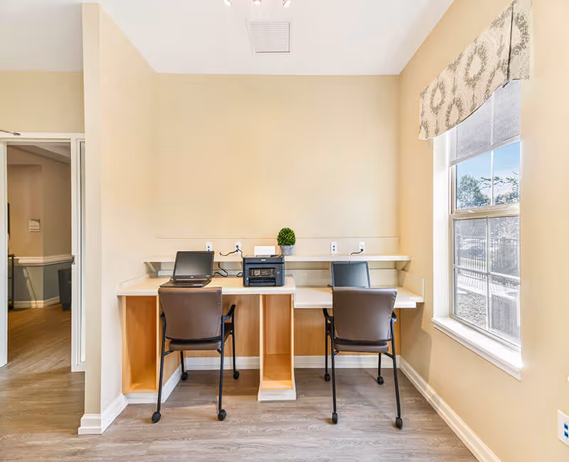 A small workspace area with two desks and chairs, each desk equipped with a laptop and a printer in the middle. The room has beige walls, a large window with a patterned valance letting in natural light, and wood flooring.