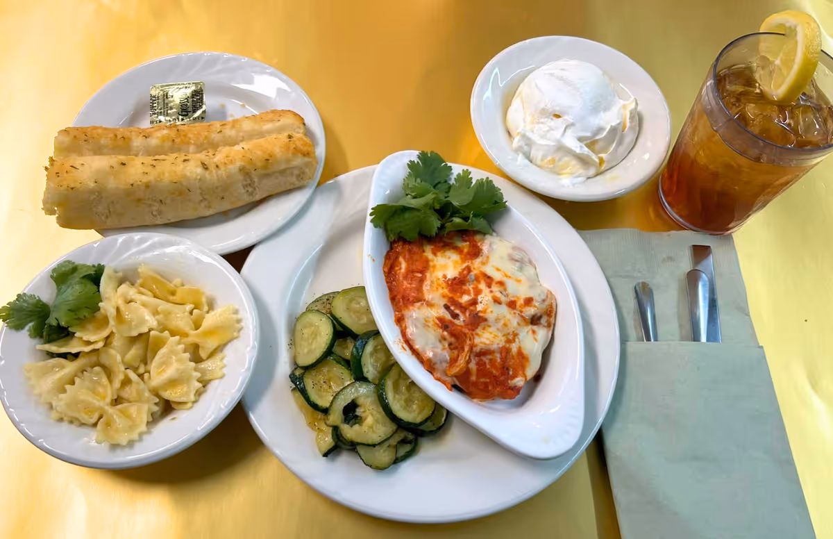 A meal served on a yellow tablecloth featuring a plate with baked dish topped with melted cheese and tomato sauce, sautéed zucchini slices, a bowl of bowtie pasta garnished with cilantro, a plate with two breadsticks and a butter packet, a bowl of whipped cream or sour cream, and a glass of iced tea with a lemon wedge. A napkin holds a fork and knife beside the plate.