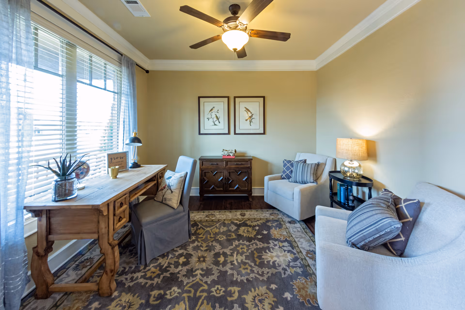 A cozy living room with two beige armchairs adorned with patterned pillows, a wooden side table with a lamp, a wooden desk with a chair and decorative items, a patterned area rug, two framed bird prints on the wall, and a ceiling fan with a light fixture.