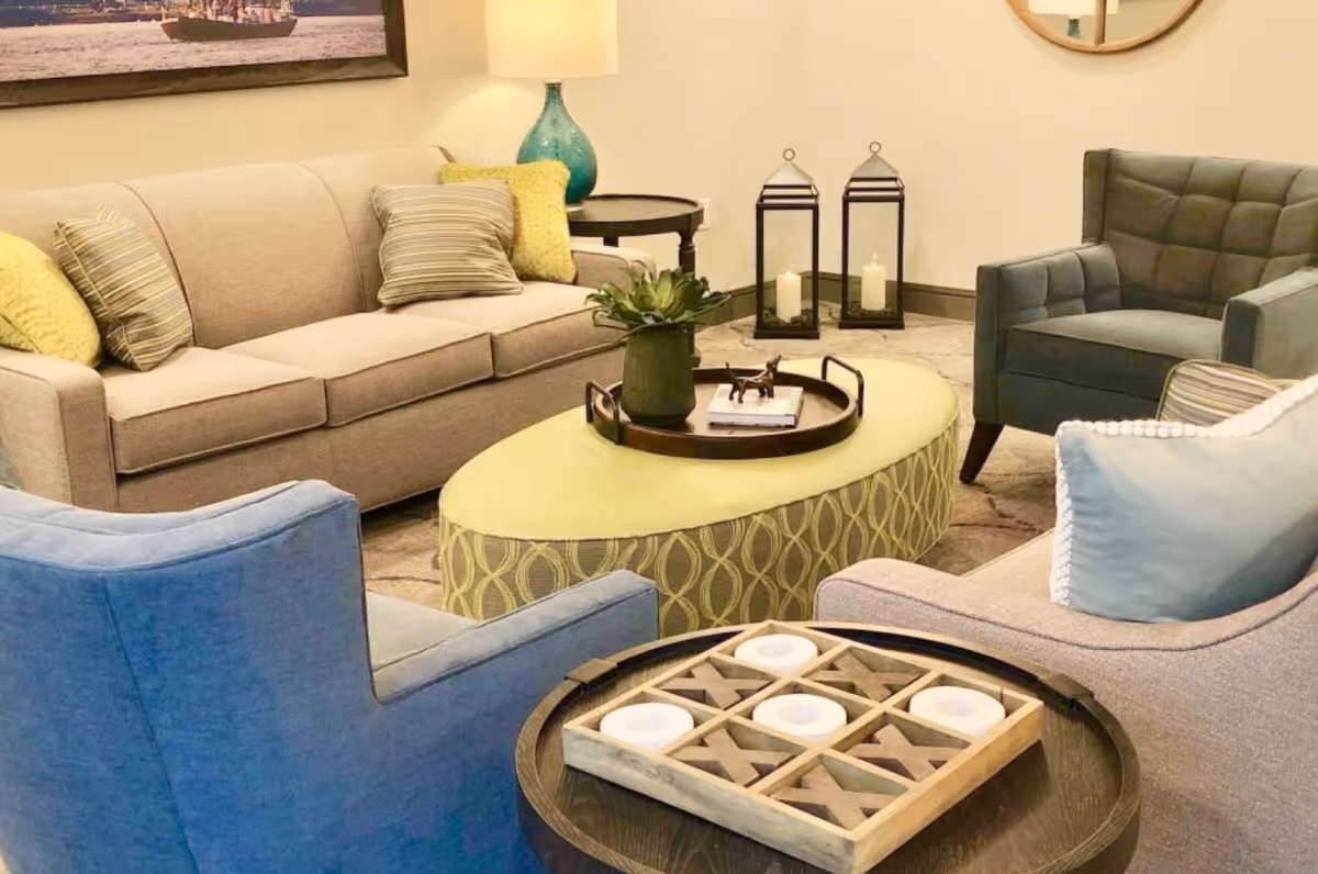 A cozy living room area with a beige sofa adorned with striped and yellow pillows, a blue armchair, and a gray armchair. In the center is an oval yellow ottoman with a decorative tray holding a plant and small items. Nearby is a round side table with a tic-tac-toe game and candles. Two large lantern candles are placed on the floor against the wall, and a table lamp with a turquoise base is on a side table behind the sofa.