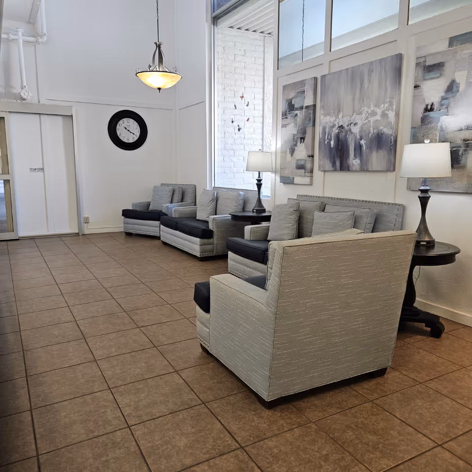 A waiting area with four cushioned armchairs arranged along a wall with two round side tables holding lamps. Three abstract paintings hang on the wall above the chairs. A large window lets in natural light. A round wall clock and a hanging light fixture are also visible. The floor is tiled.
