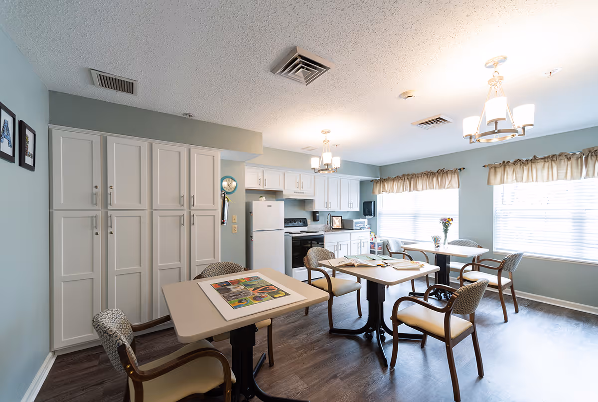 Bright communal dining area with several tables and chairs and a kitchenette in the background.