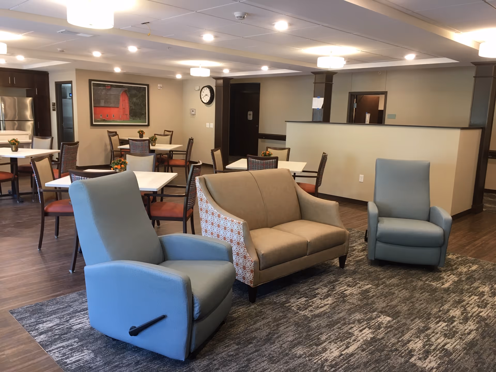 A common area in a senior living facility featuring two blue recliner chairs and a beige loveseat on a patterned rug. In the background, there are several dining tables with chairs, small flower arrangements on the tables, a large framed picture of a red barn on the wall, and a stainless steel refrigerator in the kitchen area.
