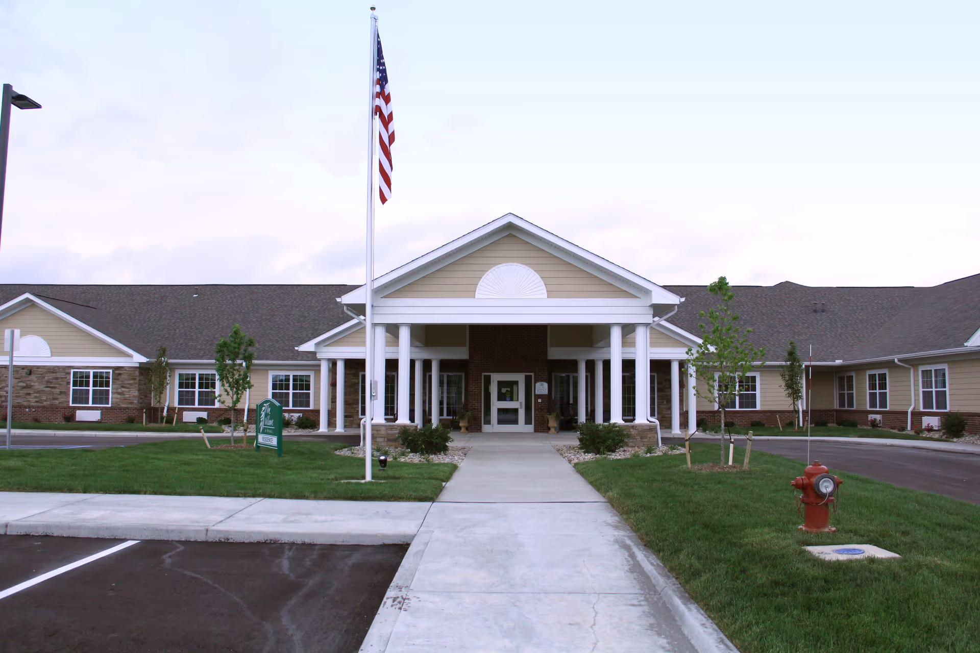 Front entrance of a single-story care facility with columns, an American flag on a flagpole, a paved walkway, and landscaped lawn.