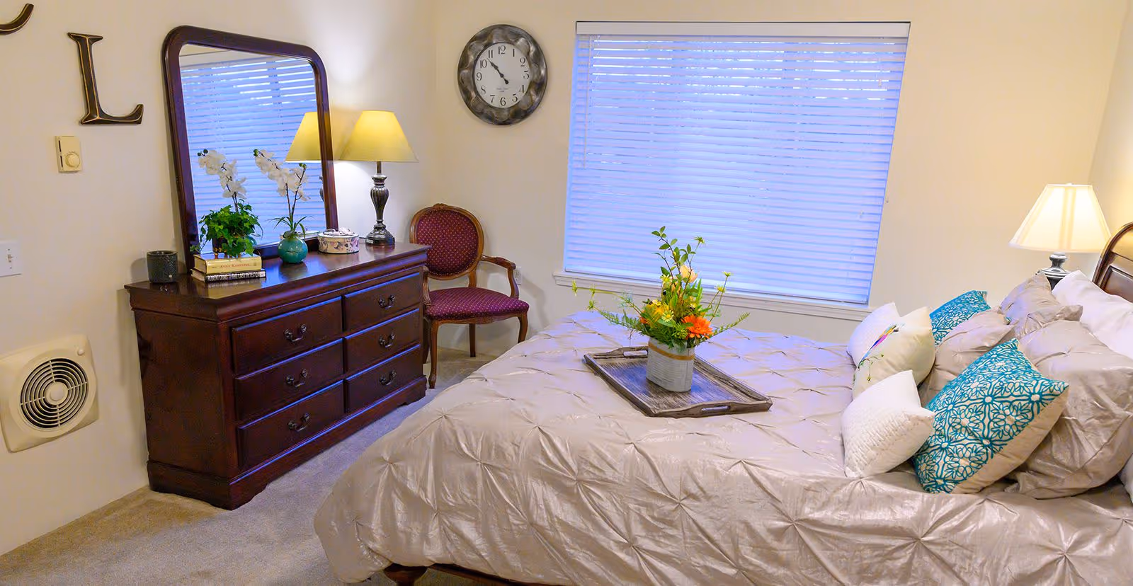 A cozy bedroom with a large bed covered in a beige comforter and multiple decorative pillows in white and blue patterns. A wooden tray with a flower arrangement sits on the bed. To the left, there is a dark wooden dresser with a large mirror, a table lamp, books, and potted plants. A red upholstered chair is placed in the corner near a window with closed blinds. A round wall clock hangs above the chair.
