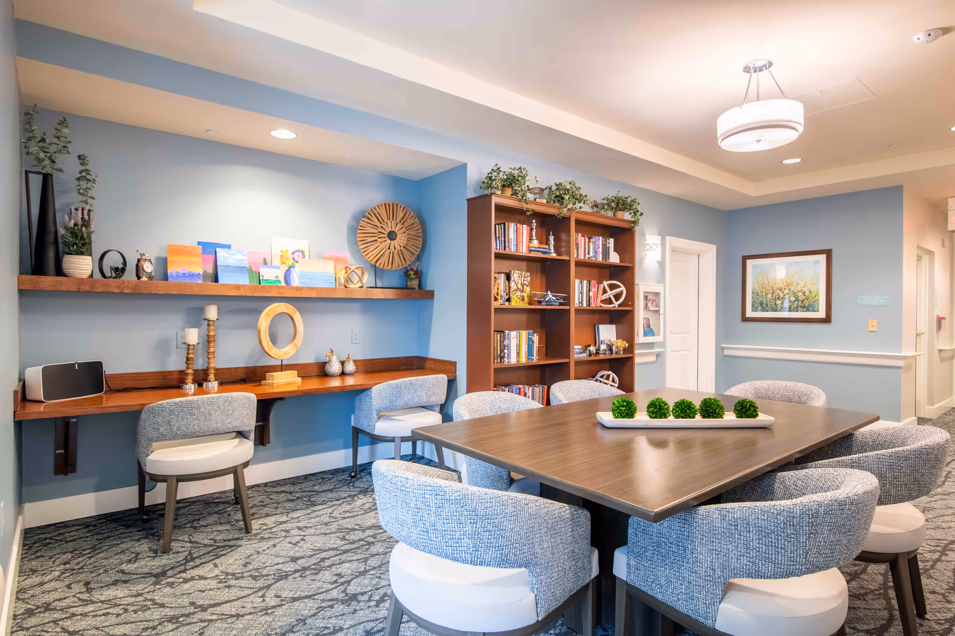 Bright communal dining and activity room with a large rectangular table surrounded by upholstered chairs, a bookshelf, and a wall-mounted desk with decorative items.