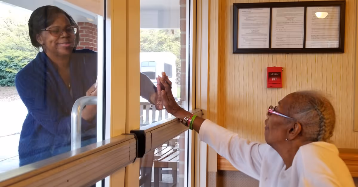 An elderly woman inside a facility reaches out to touch hands through a glass door with a woman standing outside, both smiling and connecting through the glass.