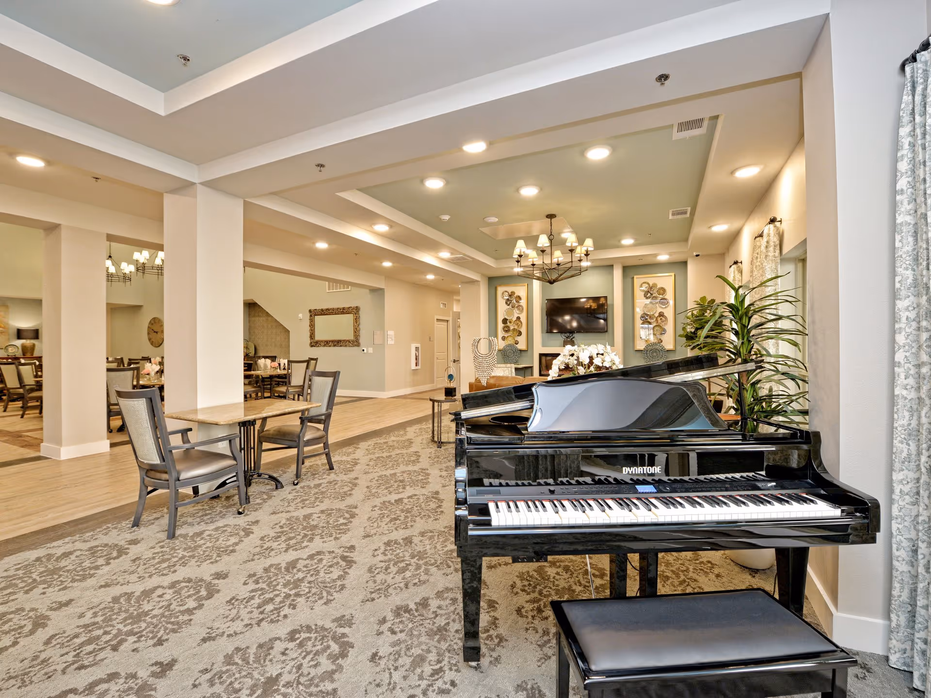 Interior view of a senior living facility lounge area featuring a black grand piano with a matching bench in the foreground. The room has patterned carpet, light-colored walls, and recessed ceiling lights. In the background, there are tables and chairs arranged for dining or socializing, decorative wall art, a flat-screen TV mounted above a fireplace, and large windows with curtains allowing natural light.